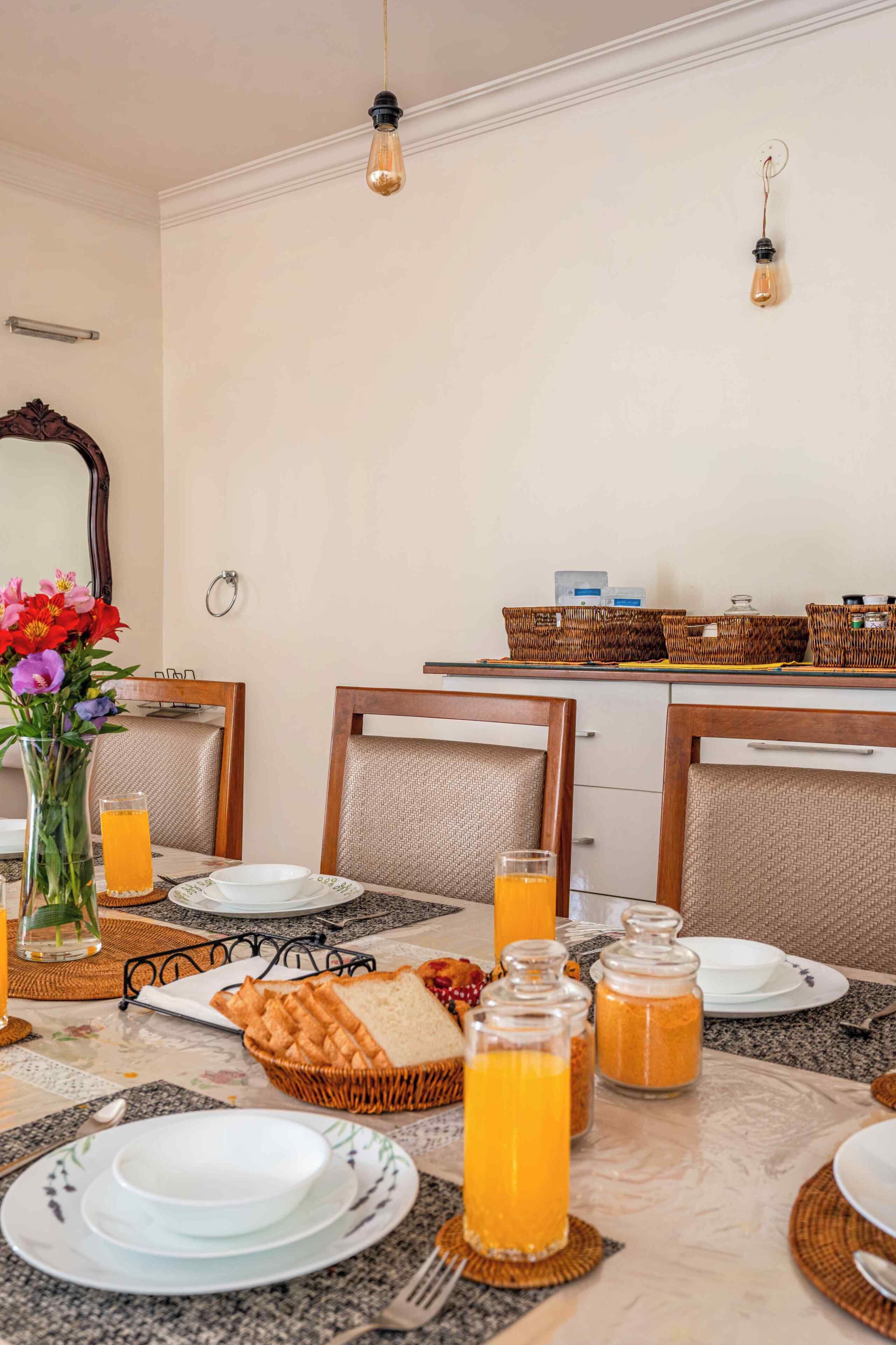 Un comedor con una mesa lista para el desayuno, con platos, vasos de zumo de naranja y cestas de pan.