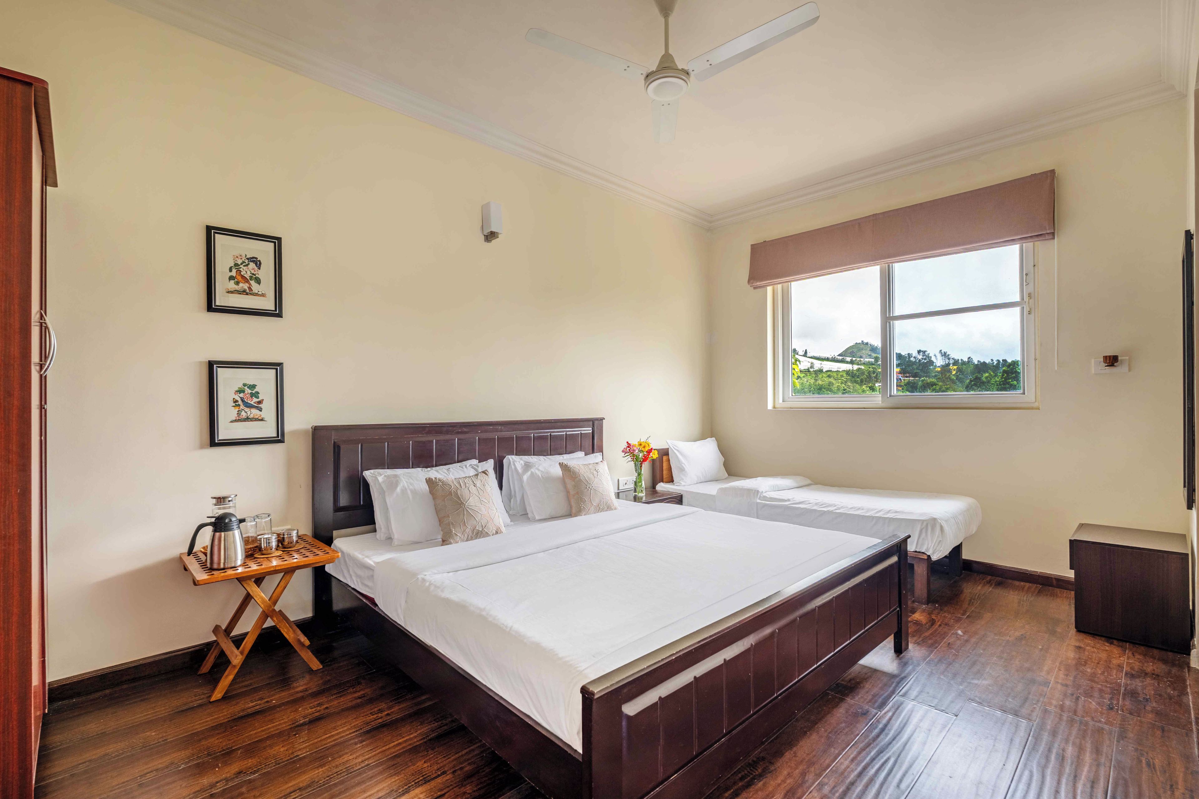 A hotel room with two beds, a wooden floor, a ceiling fan, and a window with a view of the outdoors.