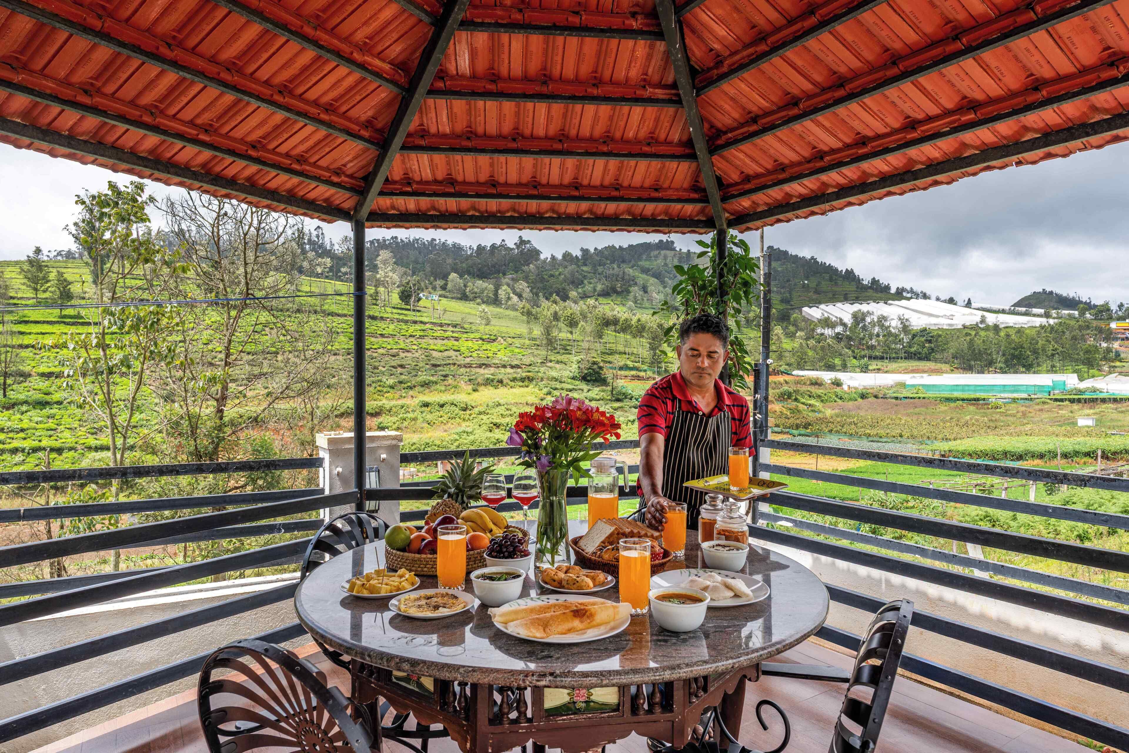 Un área de comedor al aire libre con techo, con una mesa dispuesta para una comida, rodeada de exuberante vegetación y un paisaje montañoso.