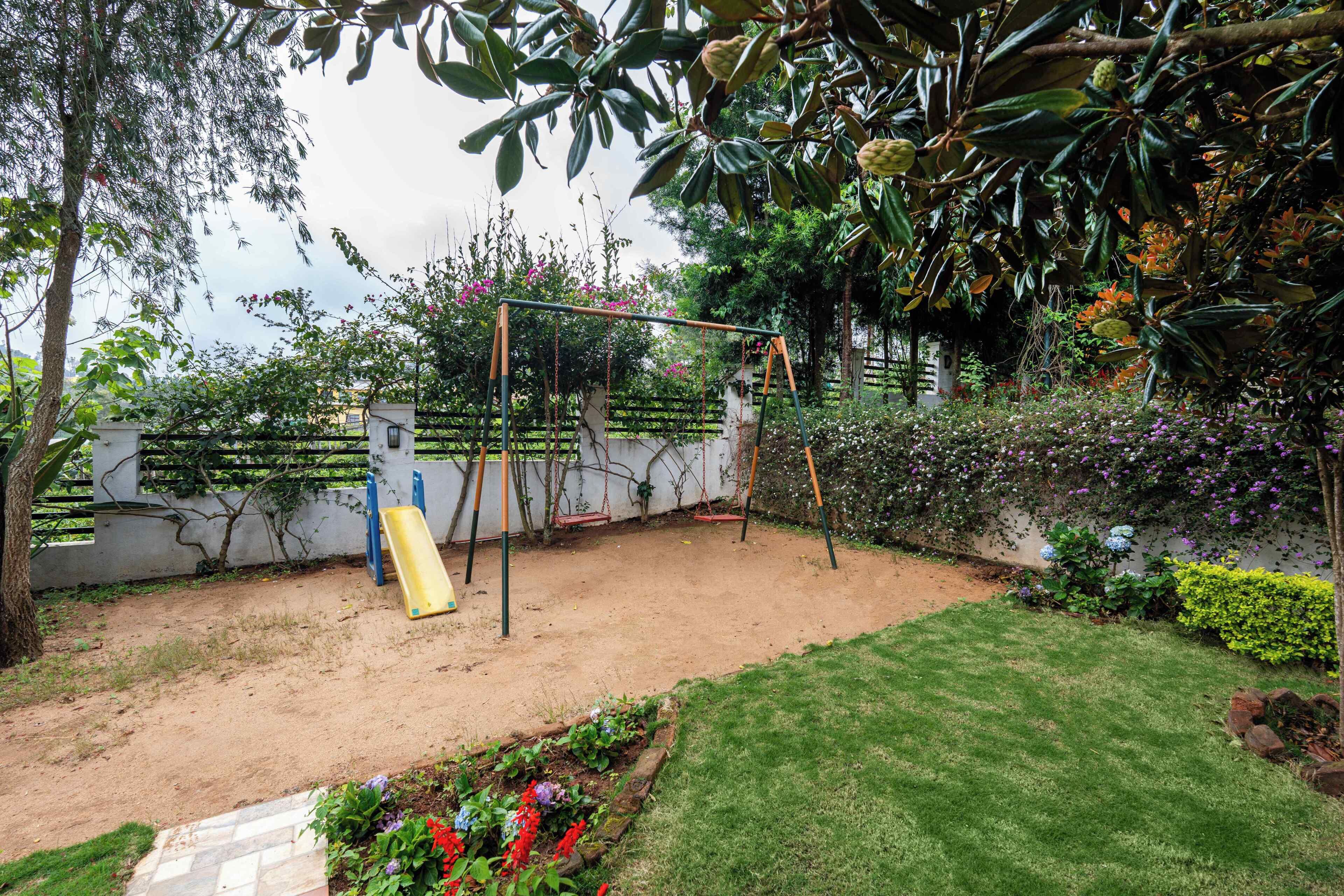 Um parquinho com balanços e escorrega, cercado por um jardim com diversas plantas e flores.