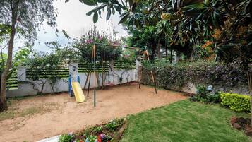 Um parquinho com balanços e escorrega, cercado por um jardim com diversas plantas e flores.