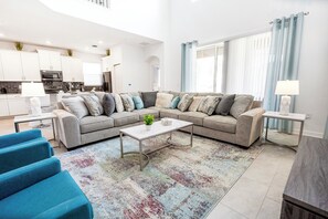 A modern living room with a sectional sofa, a coffee table, and a blue armchair. There is a kitchen in the background with white cabinets and a stove. - High Ceiling/Themed BR/SPA 22089 (Kissimmee)
