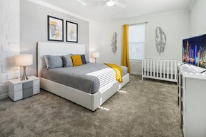 A modern bedroom with a white bed, a nightstand, a lamp, a crib, and decorative wall art. - High Ceiling/Themed BR/SPA 22089 (Kissimmee)