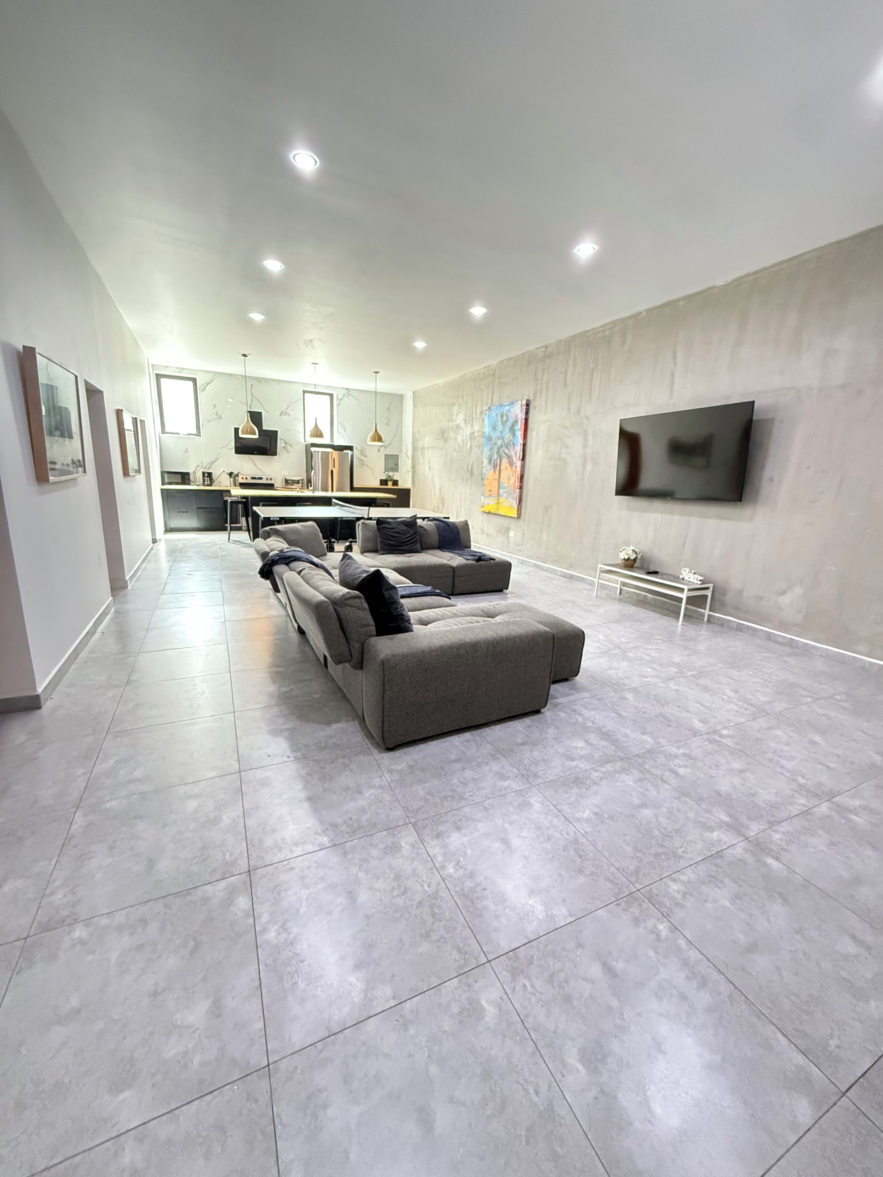 A modern living room with a grey sectional sofa, a flat-screen TV mounted on the wall, and a kitchen area in the background.