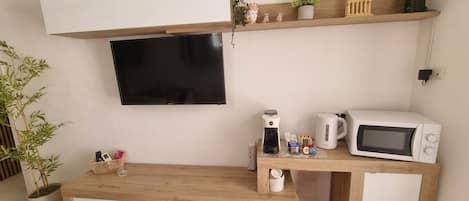 A modern living space with a flat-screen TV mounted on the wall, a wooden coffee table with storage, a microwave, and a kettle on a wooden countertop.