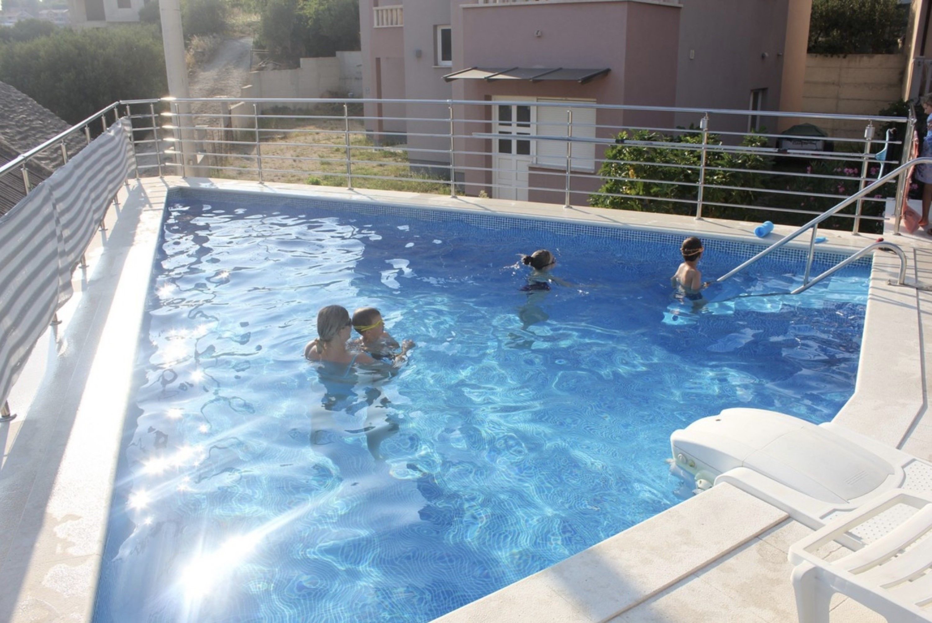 A swimming pool with three people in the water, surrounded by a metal railing and a white lounge chair.