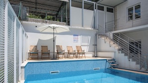 Una piscina con borde de azulejos, sillas de lounge de madera, una zona cubierta con techo metálico y una escalera que conduce a un nivel superior.