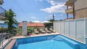 Una piscina con sillones de descanso y vistas a un edificio con techo de tejas rojas.