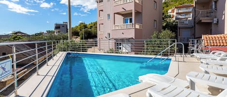 A swimming pool with lounge chairs, a metal railing, and a building with balconies in the background.