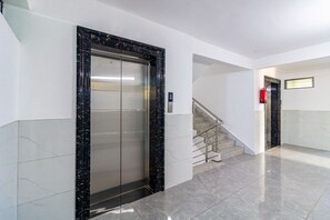 Un ascensor modern amb marc de marbre, una escala amb baranes metàl·liques i un extintor fixat a la paret.