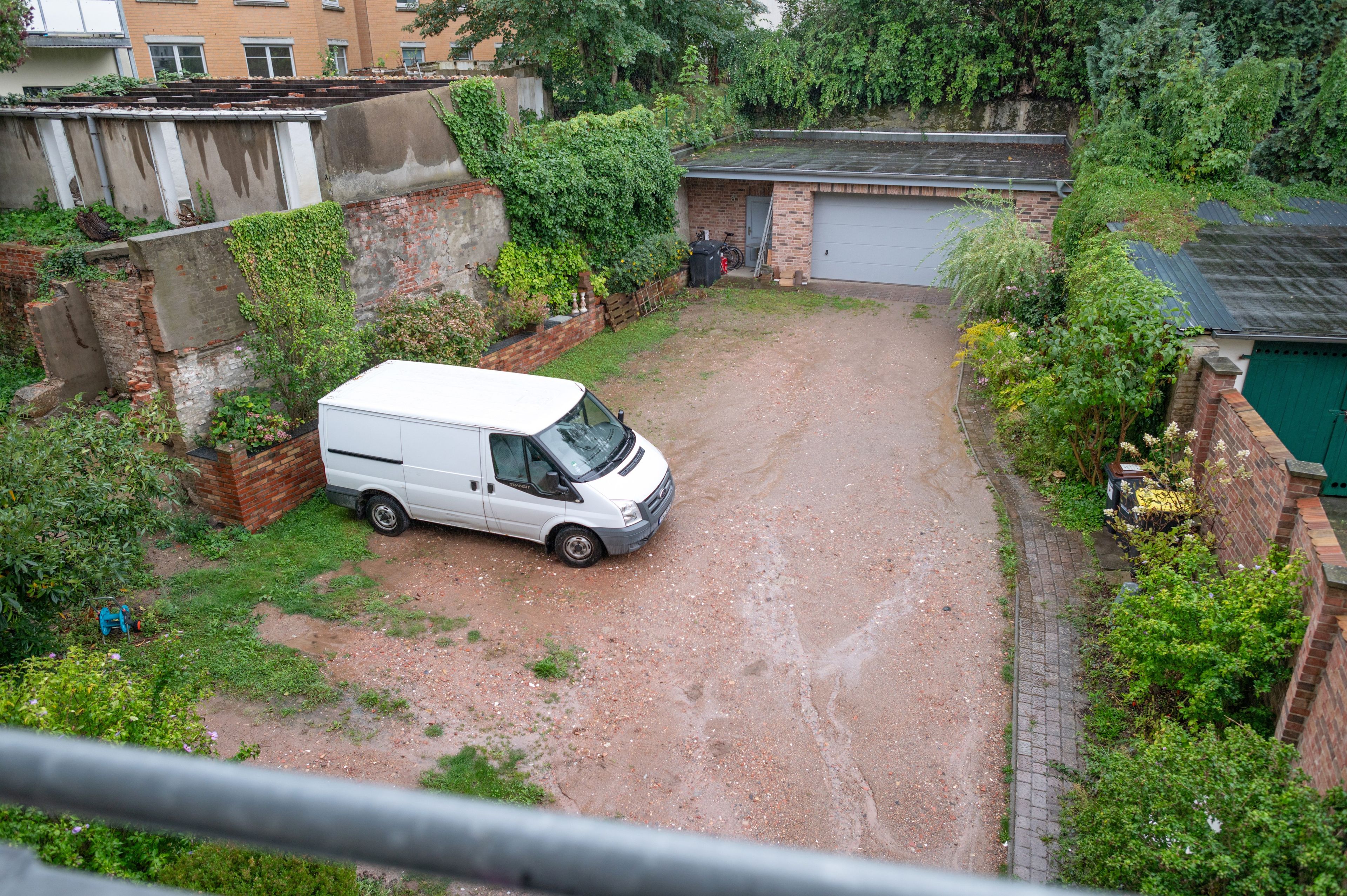 Una camioneta blanca estacionada en un camino de grava con un muro de ladrillo y vegetación a un lado, y un garaje al otro.