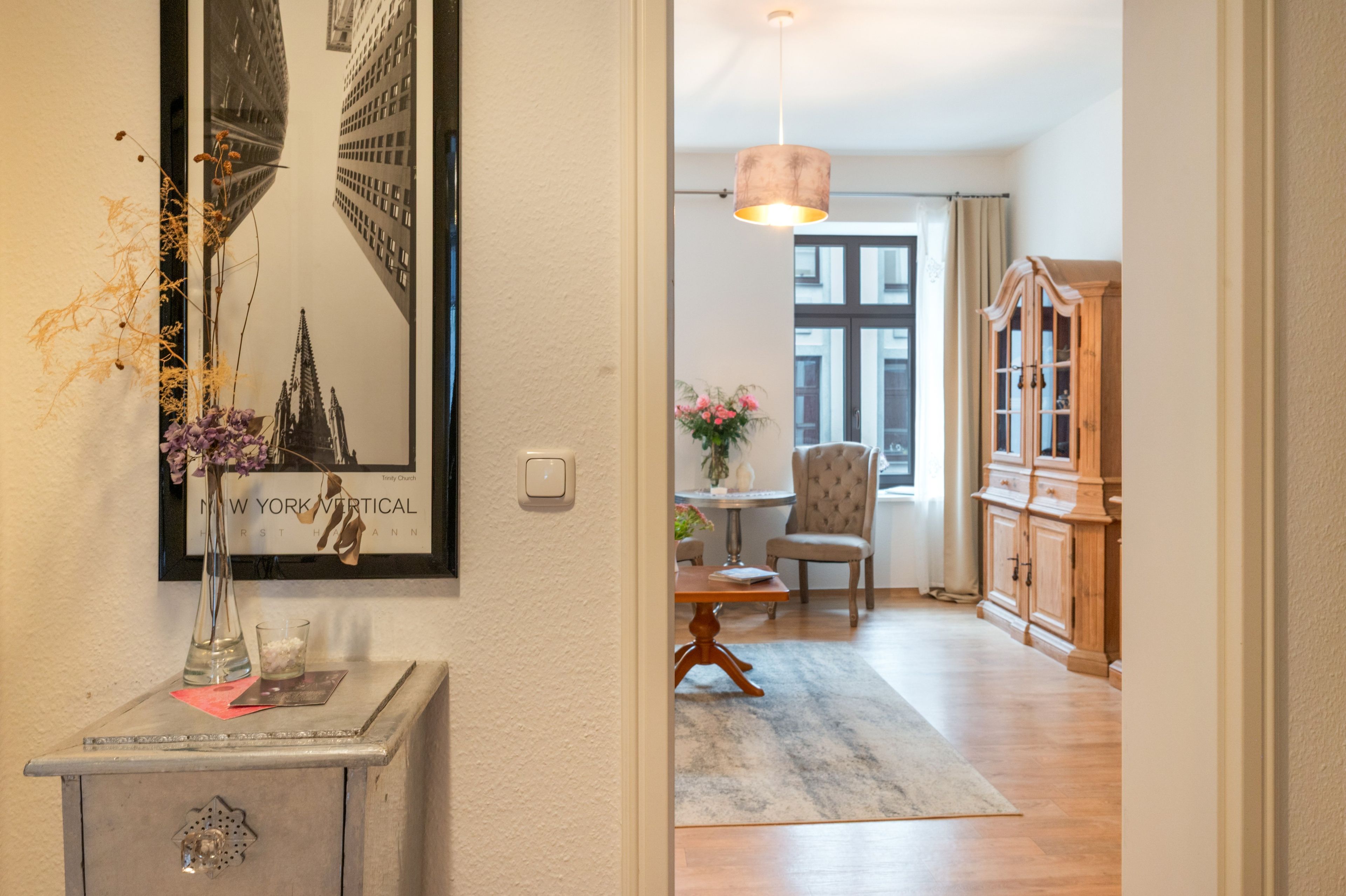 A room with a framed picture of a building, a small table with a chair, a cabinet, and a hanging light fixture.