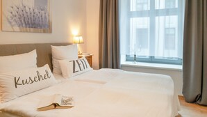 A neatly made bed with pillows labeled 'Kuschel' and 'Zeit' in a hotel room with a large window and a lamp on the nightstand.