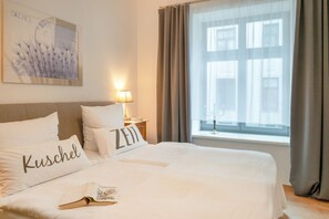 A neatly made bed with pillows labeled 'Kuschel' and 'Zeit' in a hotel room with a large window and a lamp on the nightstand.