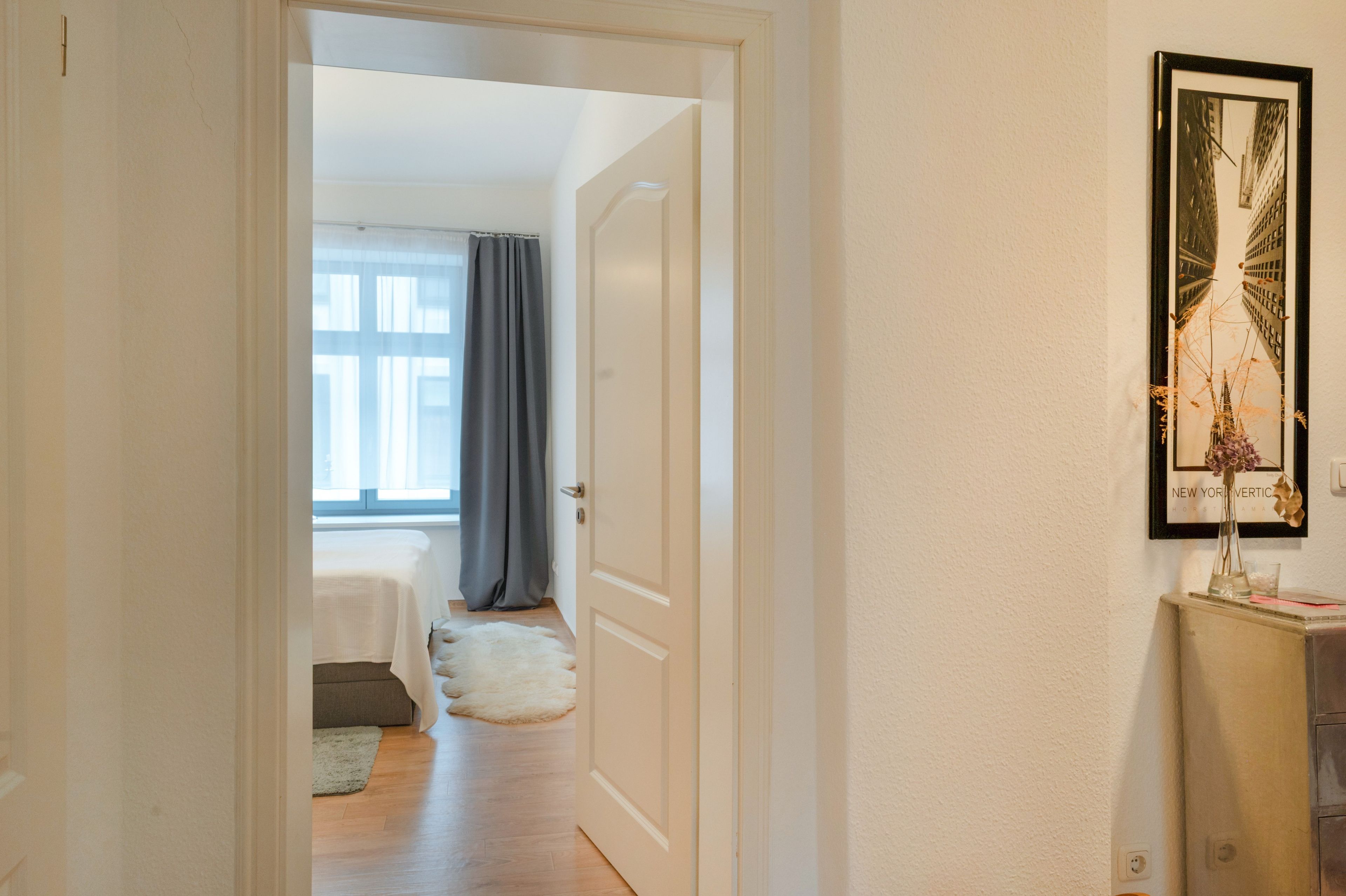 Un pasillo con una puerta que da a un dormitorio con cama y ventana con cortinas.