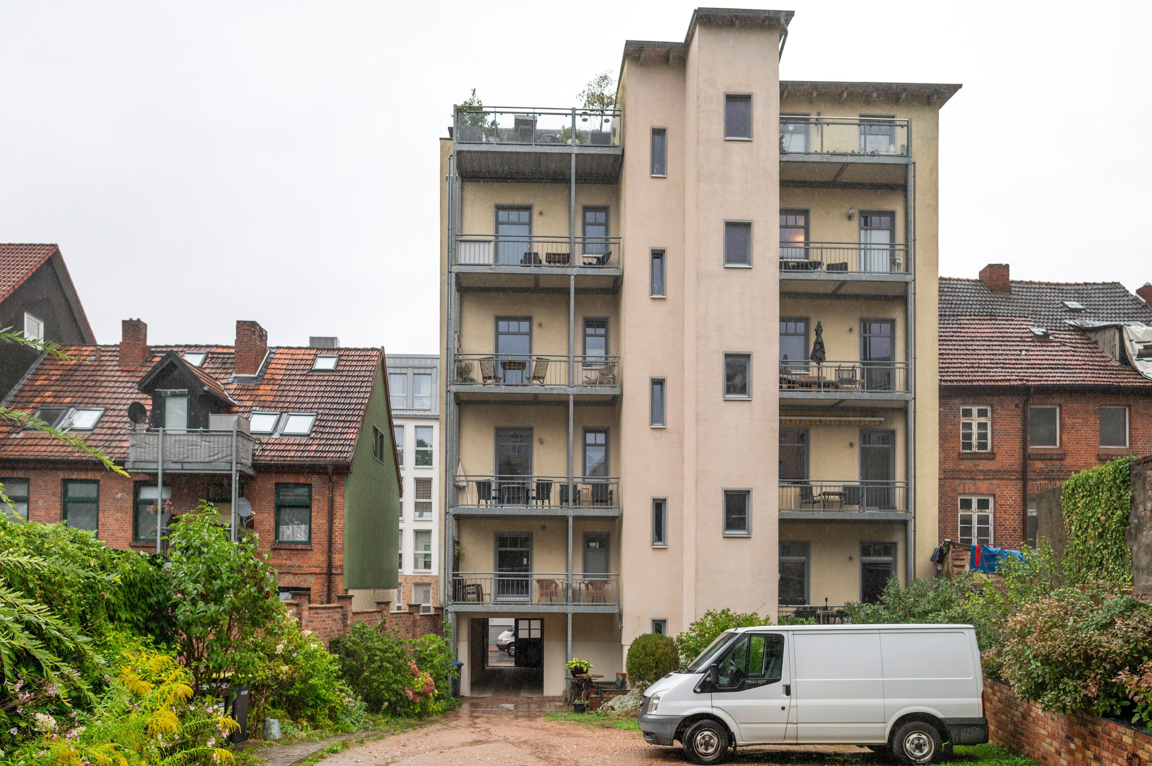 Un edificio residencial de varios pisos con balcones, una camioneta blanca estacionada frente a él y un área de jardín con diversas plantas.
