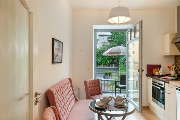 A compact kitchen with modern appliances, a dining area with a table set for tea, and a view of a balcony with greenery.