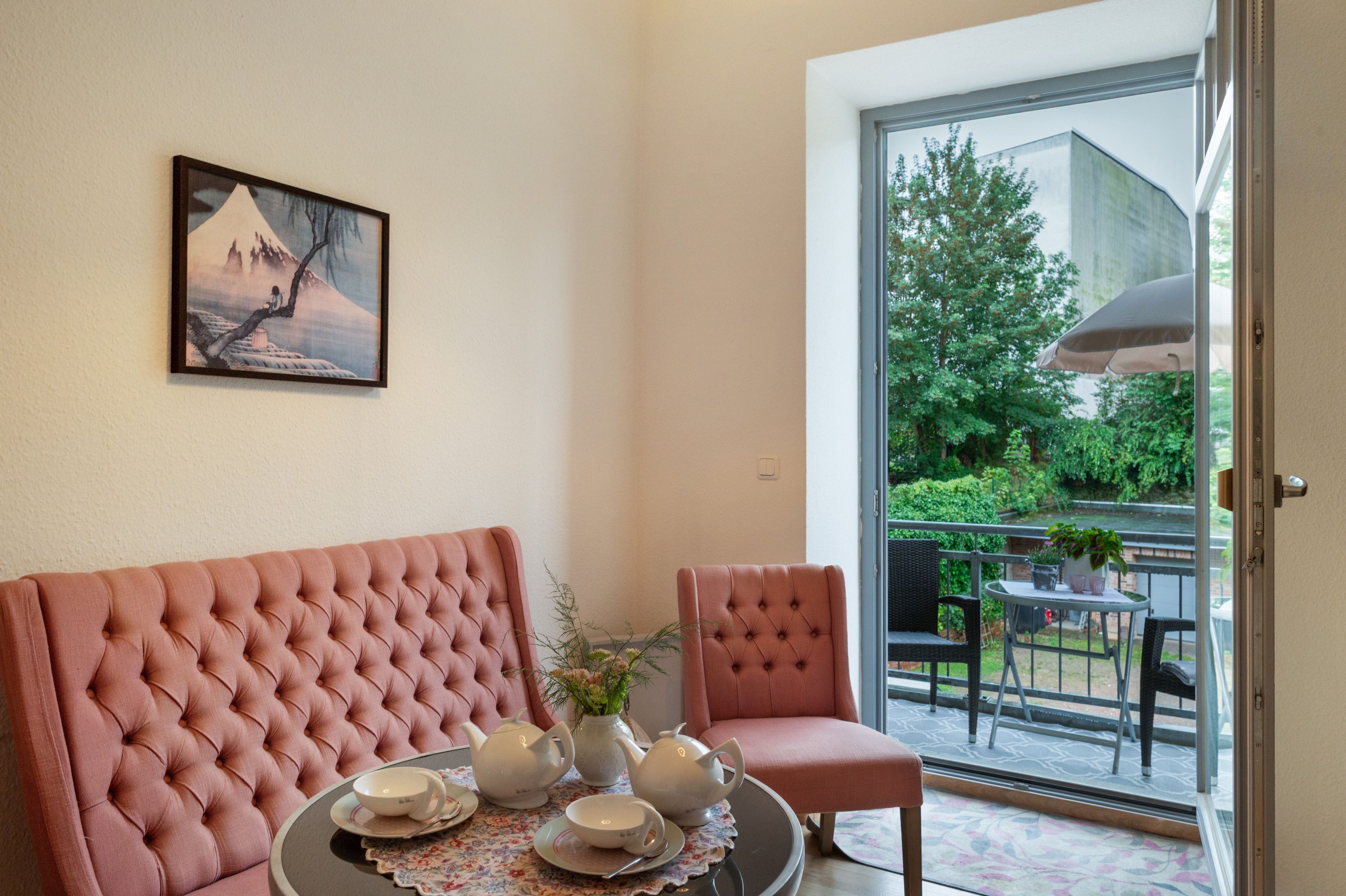 A room with a pink upholstered sofa, a dining table set with teapots and cups, a painting on the wall, and a view of greenery through a glass door.