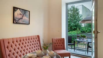 A room with a pink upholstered sofa, a dining table set with teapots and cups, a painting on the wall, and a view of greenery through a glass door.