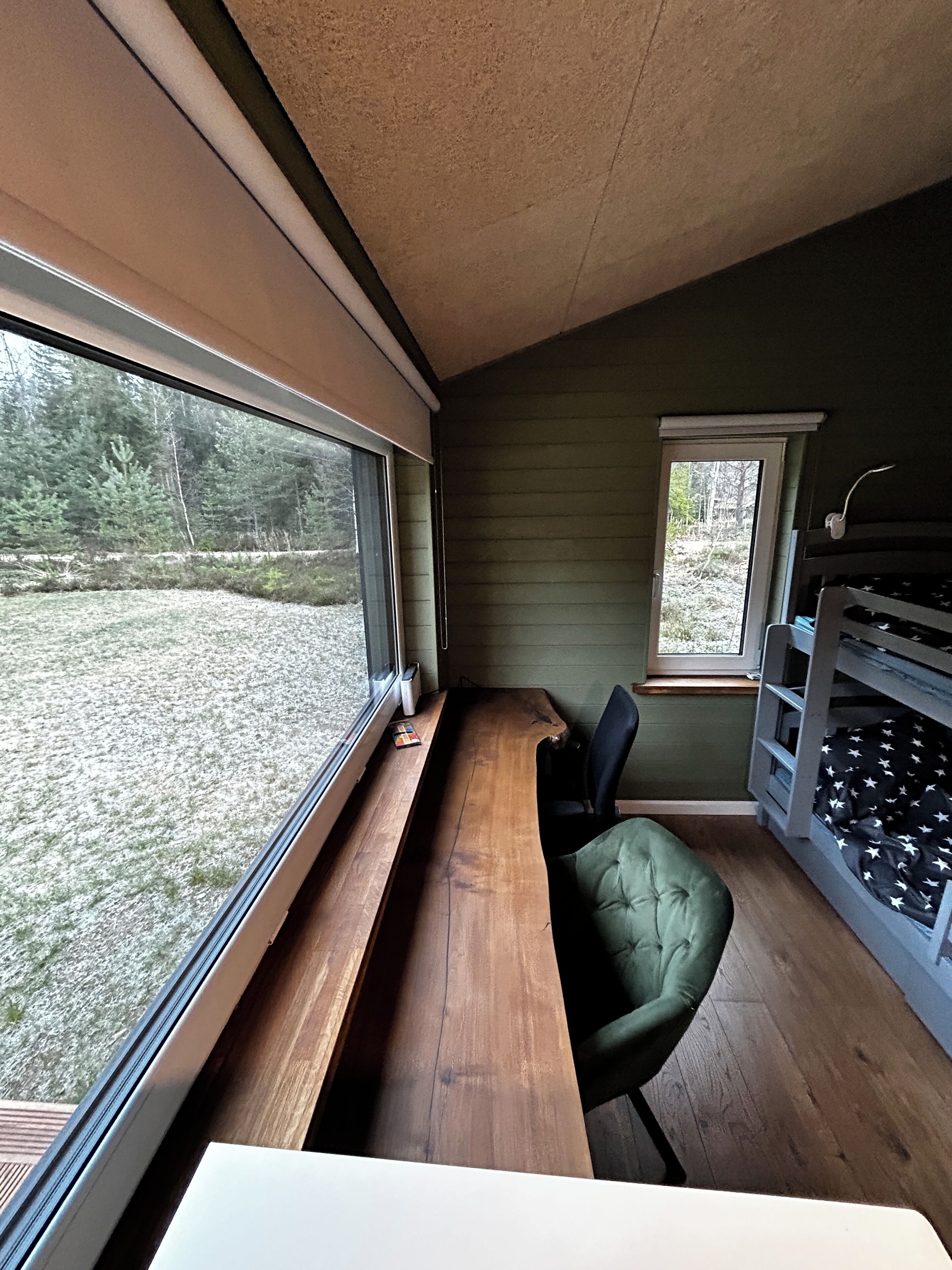 Une pièce avec un banc en bois, un fauteuil vert rembourré, des lits superposés et de grandes fenêtres donnant sur un espace verdoyant.
