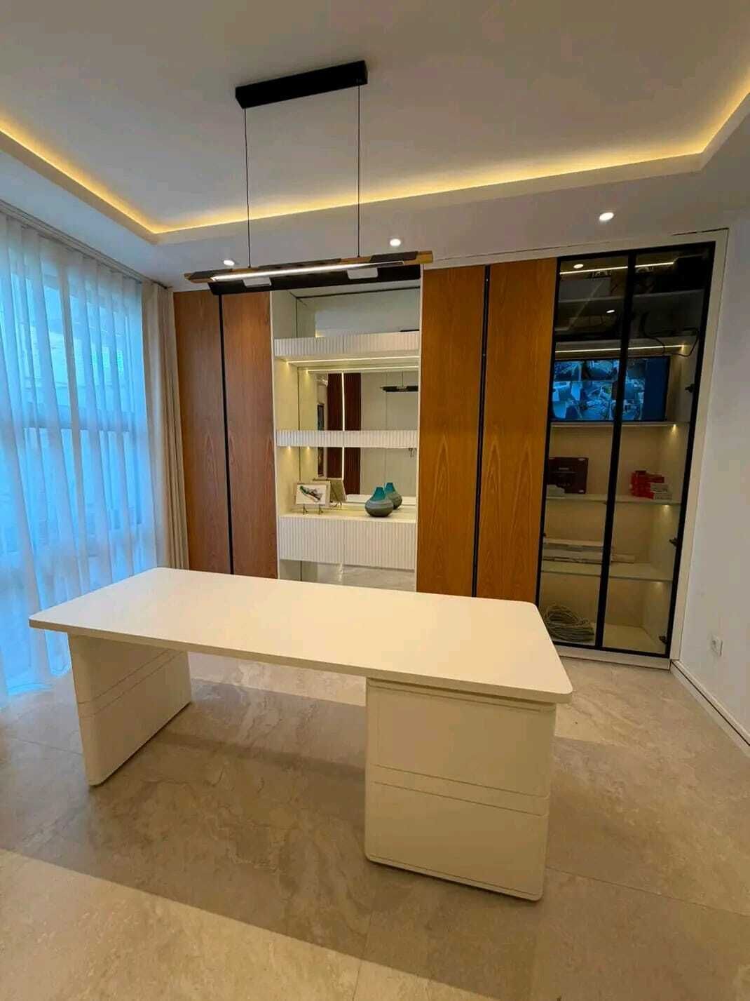 A modern room with a large white desk, a built-in wardrobe with mirrors, and a glass-fronted storage unit.