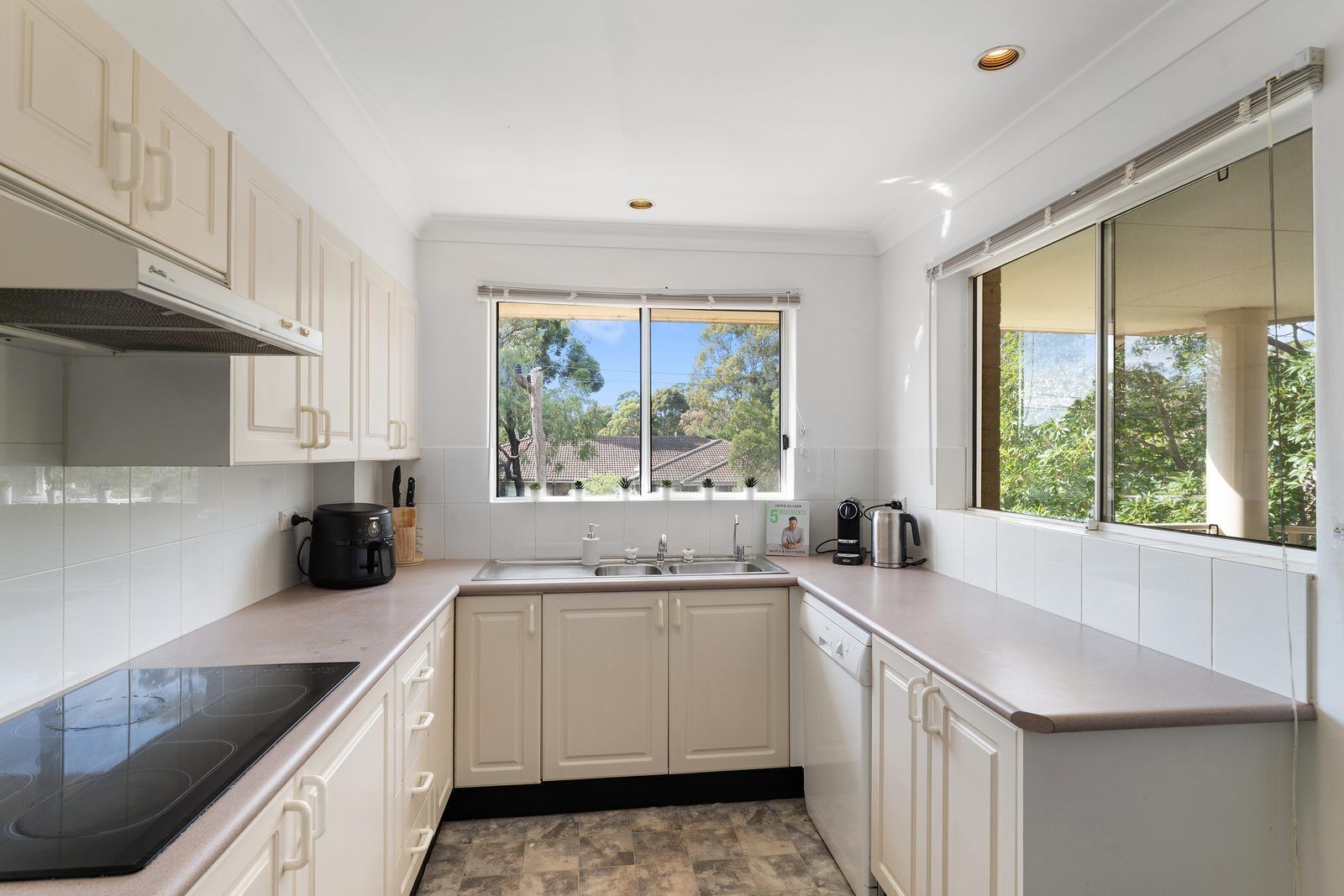 A modern kitchen with white cabinetry, a black stovetop, a window with a view of trees, and a coffee maker on the counter.