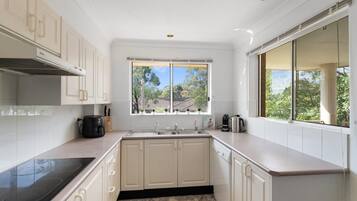 A modern kitchen with white cabinetry, a black stovetop, a window with a view of trees, and a coffee maker on the counter.