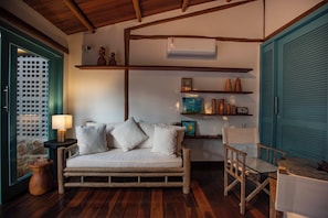 A cozy room with a wooden floor, a sofa with cushions, a small table with a lamp, and a chair. There are shelves with decorative items and a window with a view. - POUSADA VEREDAS NORONHA (Fernando de Noronha)
