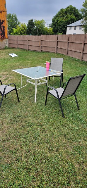 A backyard with a metal table and two chairs, a wooden fence, and a house in the background.