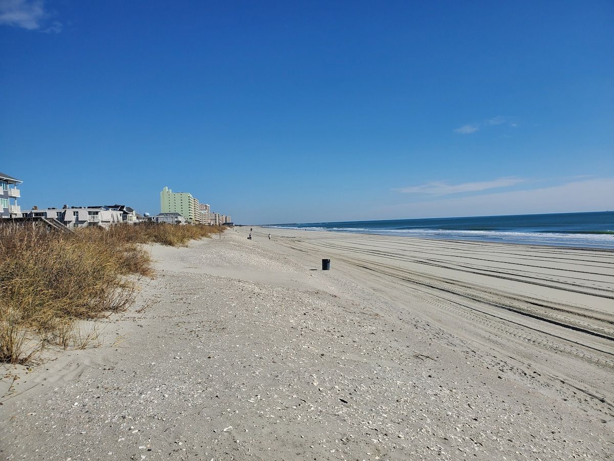 A beach with a clear sky, a sandy path leading to the water, and buildings in the background.