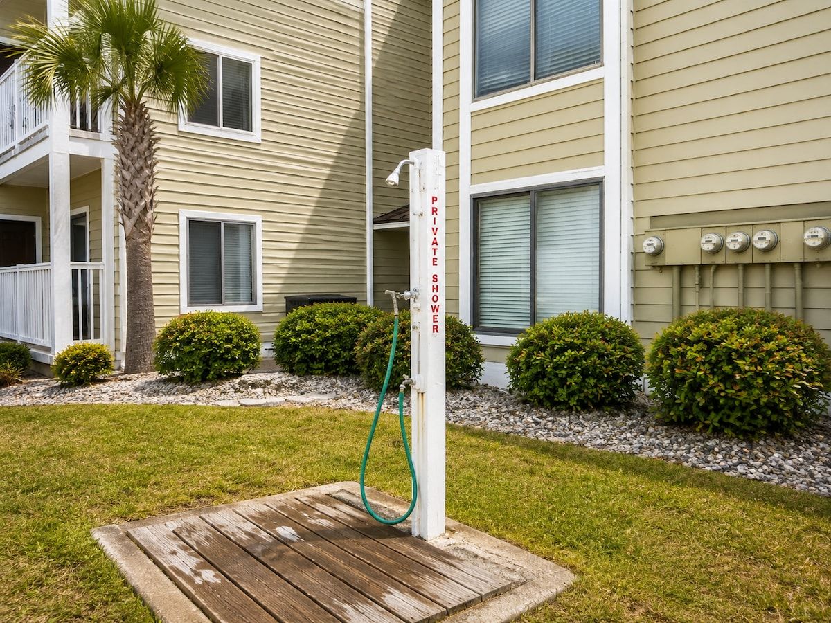 A private shower with a hose, a palm tree, and a wooden deck in front of a building with multiple windows and a balcony.