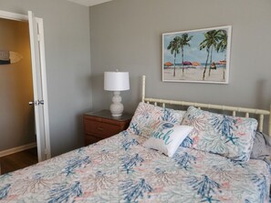Een slaapkamer met een bed, een nachtkastje, een lamp en een schilderij met een strandthema aan de muur.