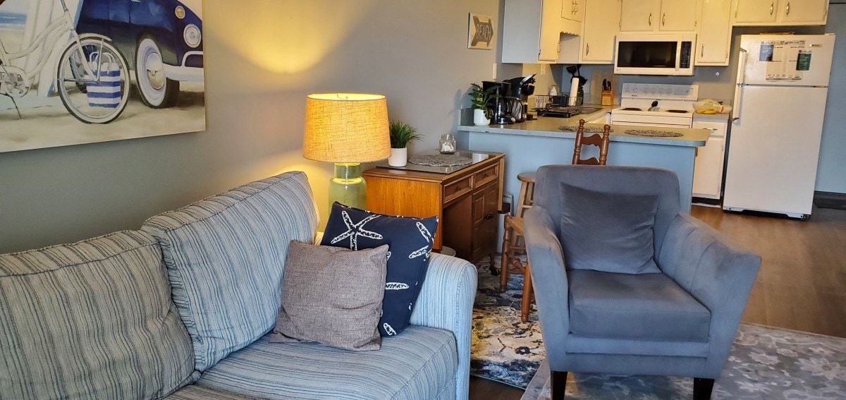 Een woonkamer met een bank, fauteuil en een lamp. Achtergrond is een keuken met kasten en apparatuur.