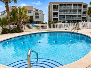Une piscine entourée d’une clôture blanche, avec des palmiers et un immeuble à plusieurs étages en arrière-plan.