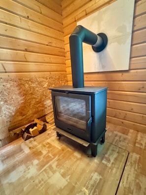 A wood-burning stove in a log cabin with a chimney, a window, and a small amount of wood on the floor. - Holiday Home 'Base-Butze Hohegeiß Log Cabin Harz' with Wi-Fi and Air Conditioning (Braunlage)