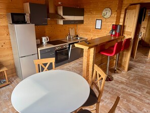 A compact kitchen with a refrigerator, microwave, stove, and sink. There is a wooden table with two chairs and a red bar stool. - Holiday Home 'Base-Butze Hohegeiß Log Cabin Harz' with Wi-Fi and Air Conditioning (Braunlage)