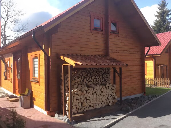 Una cabaña de madera con un porche cubierto, leña apilada y un ventanal pequeño.