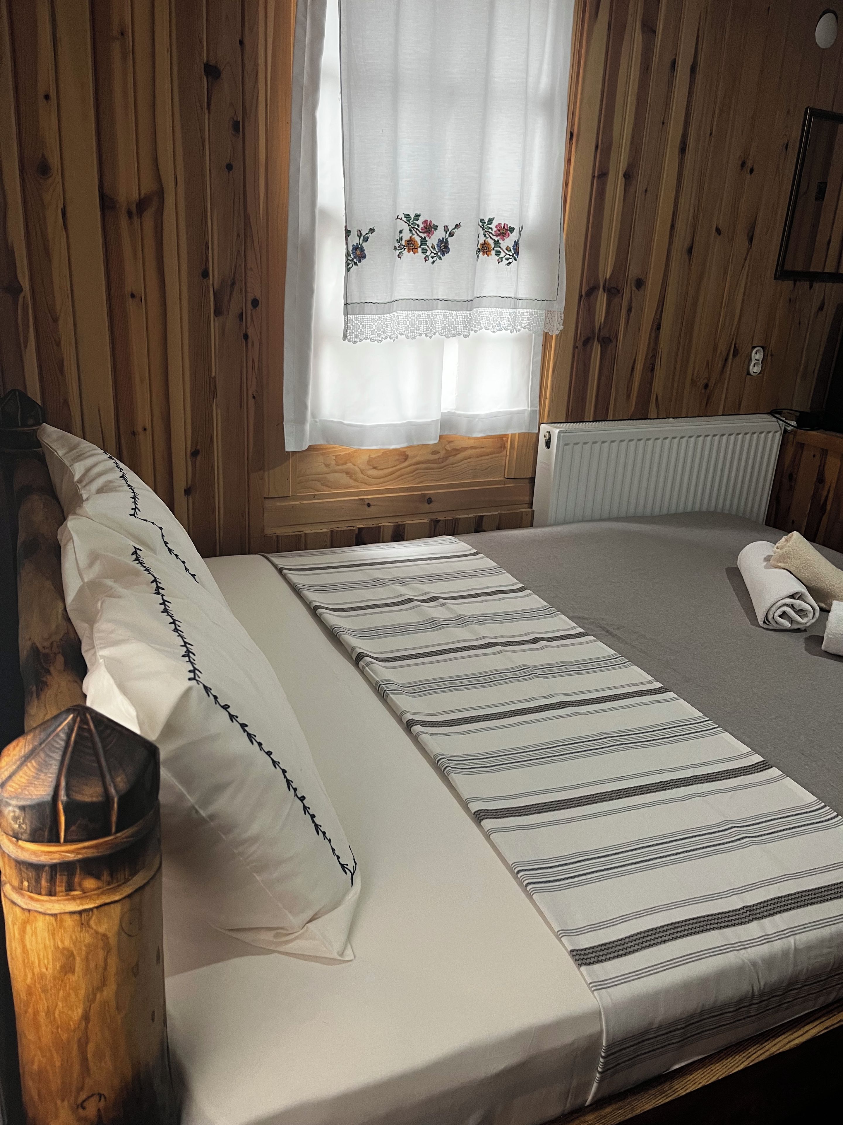 Habitación con paneles de madera, una cama, almohadas, una manta, un radiador y una ventana con cortina de motivos florales.