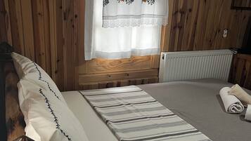 Habitación con paneles de madera, una cama, almohadas, una manta, un radiador y una ventana con cortina de motivos florales.