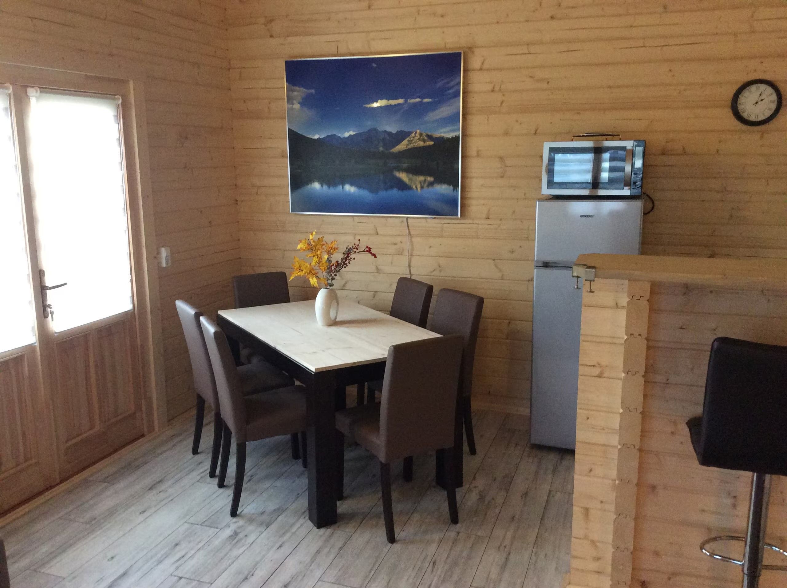 A dining area with a table, chairs, a vase with flowers, a refrigerator, and a clock on the wall.