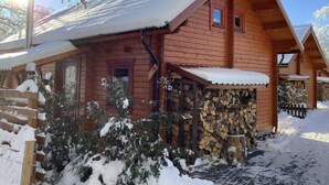 A wooden cabin with a snow-covered roof, a wooden porch, and a stack of firewood.