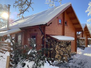 A wooden cabin with a snow-covered roof, a wooden porch, and a stack of firewood.