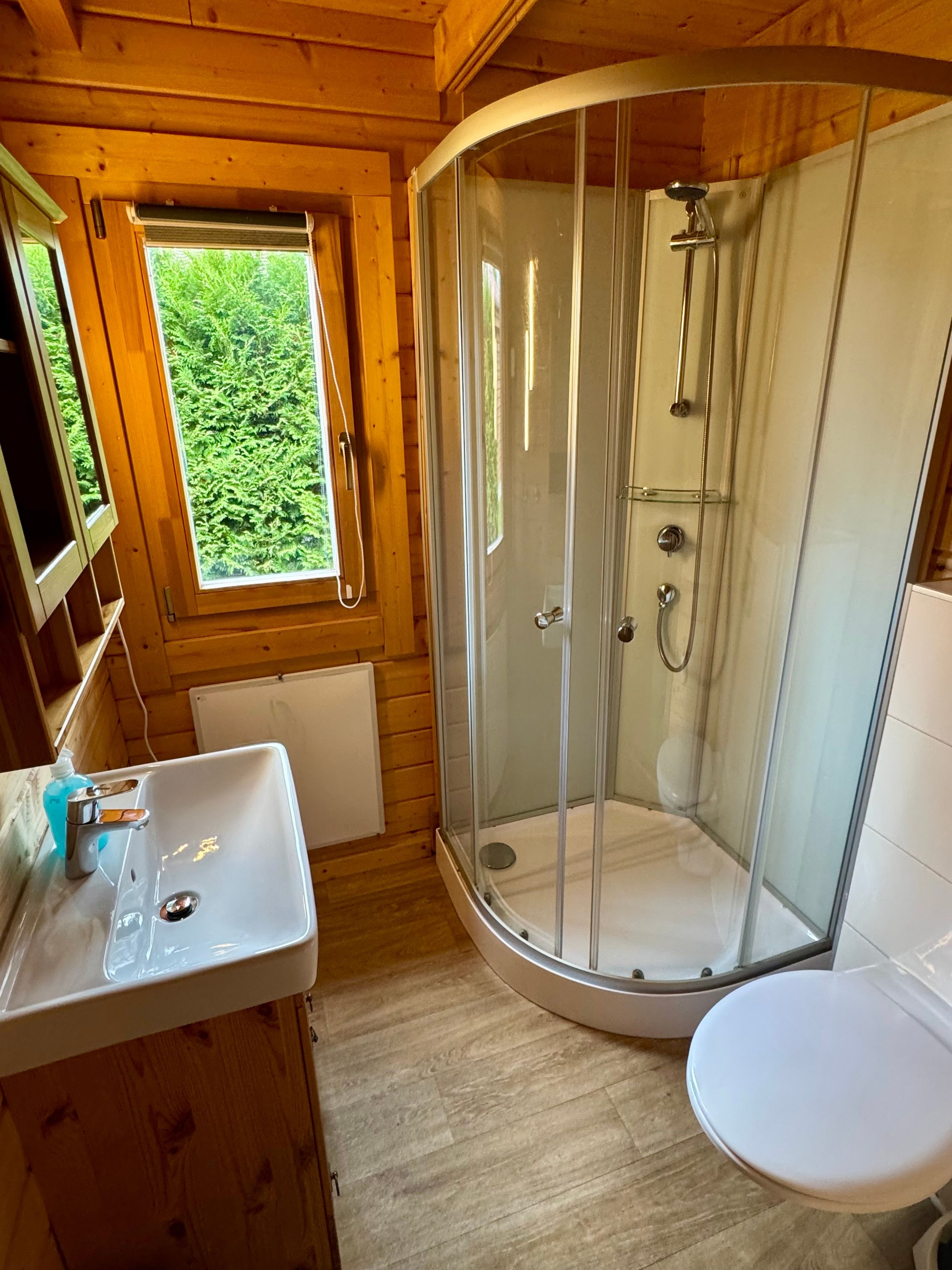 A wooden cabin bathroom with a glass-enclosed shower, a white sink, and a window with a view of greenery.
