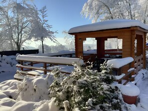 A wooden cabin with a bench and snow-covered plants in a snowy outdoor setting.