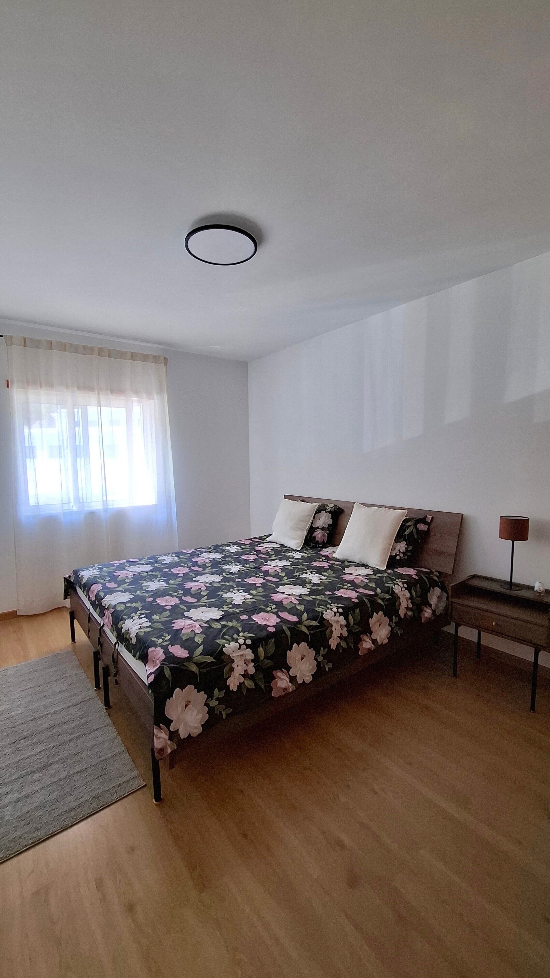 Un dormitorio con cama, mesita de noche, lámpara y ventana con cortinas.