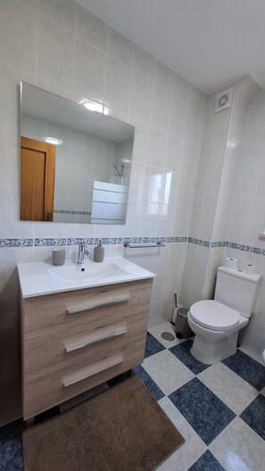 A bathroom with a white sink, wooden vanity, a mirror above the sink, a toilet, and patterned floor tiles.