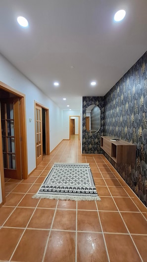 A hallway with tiled flooring, wooden doors, a patterned rug, recessed ceiling lights, and a decorative wall with a mirror.