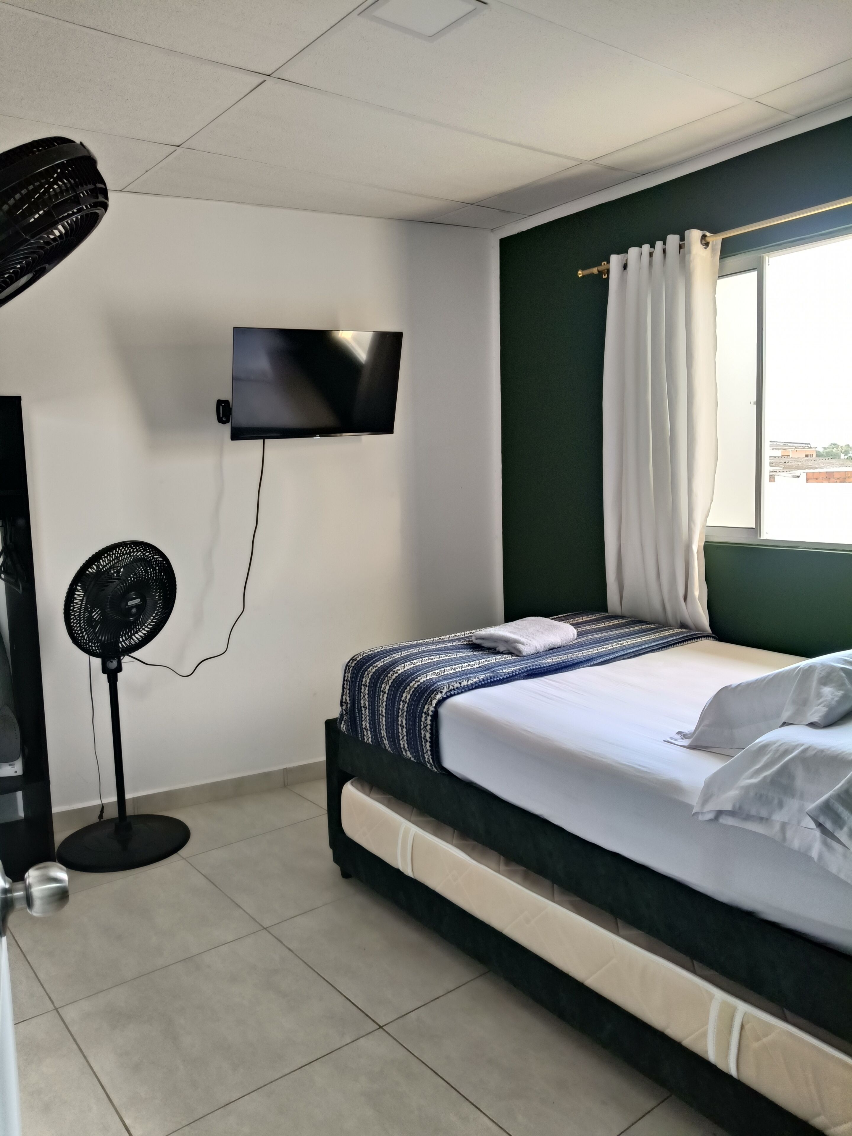 Una habitación de hotel con una cama, un televisor montado en la pared, un ventilador de pie y una ventana con cortinas.