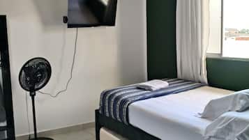 Una habitación de hotel con una cama, un televisor montado en la pared, un ventilador de pie y una ventana con cortinas.