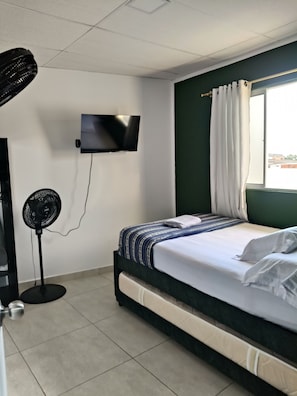 Een hotelkamer met een bed, een aan de muur bevestigde televisie, een ventilator en een raam met gordijnen.